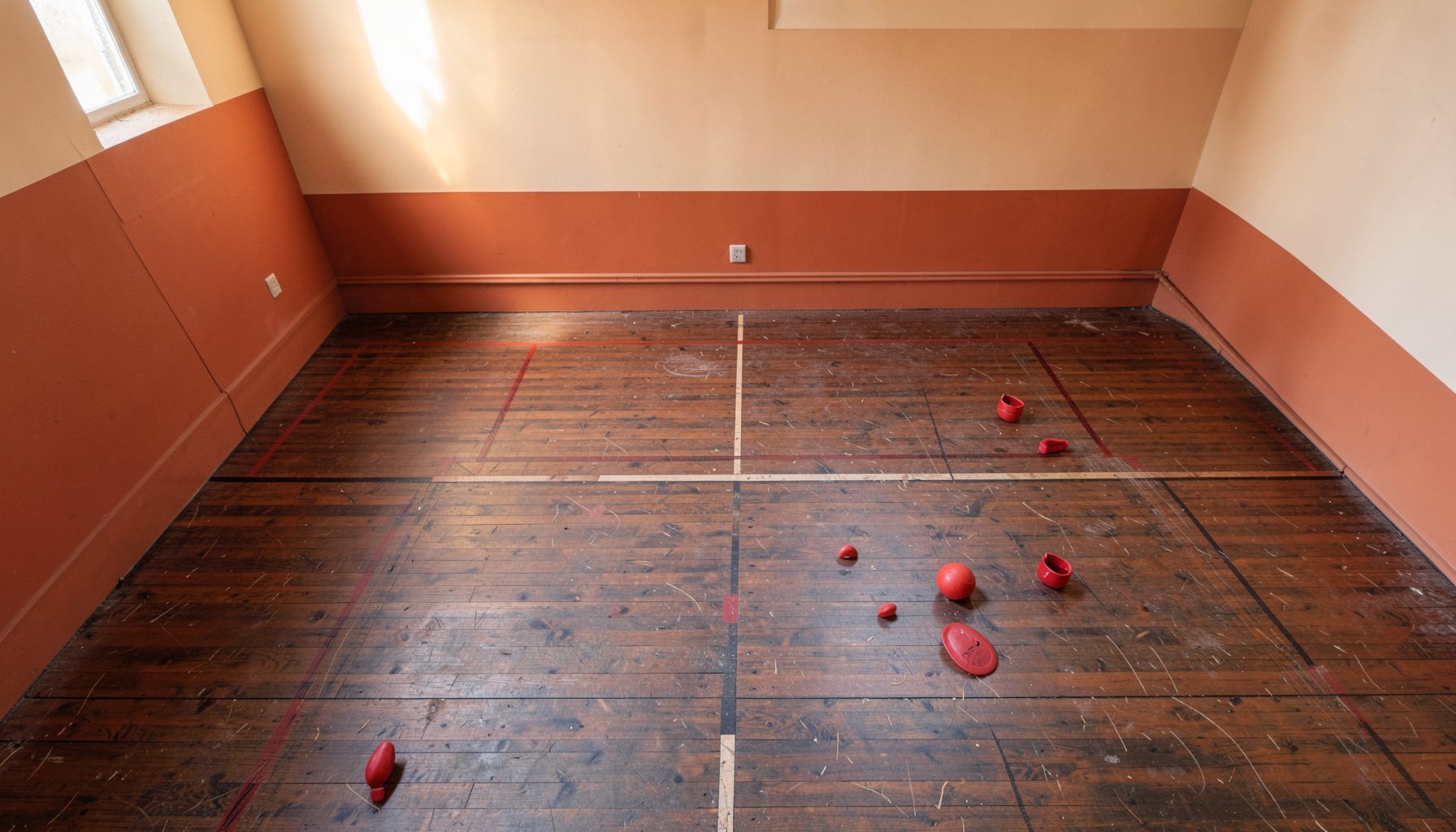 Wide-angle view of an empty Rugby Fives court with warm terracotta and cream walls, dark wooden floorboards, and soft natural light streaming from above
