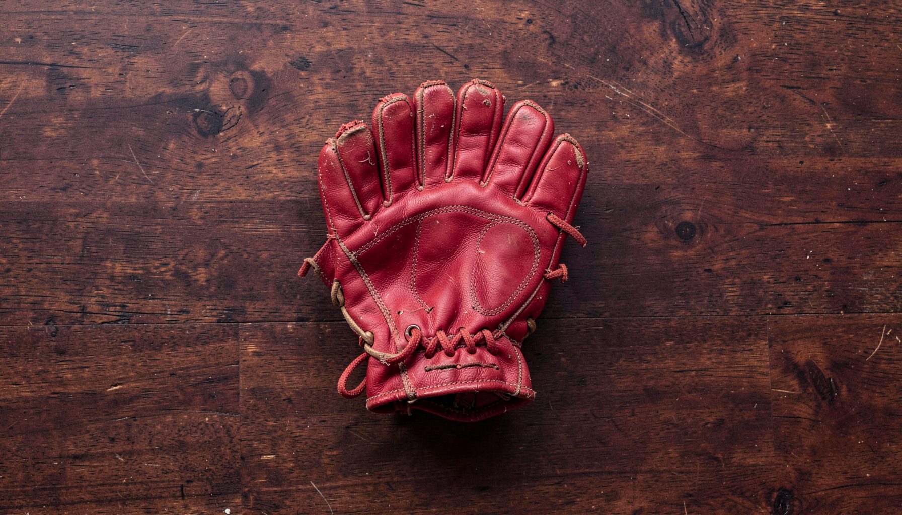 Warm-toned overhead flat-lay of a worn leather Rugby Fives glove resting on a dark wooden court floor, with a single white ball nearby; deep maroon and cream tones dominate, with soft natural light casting gentle shadows.