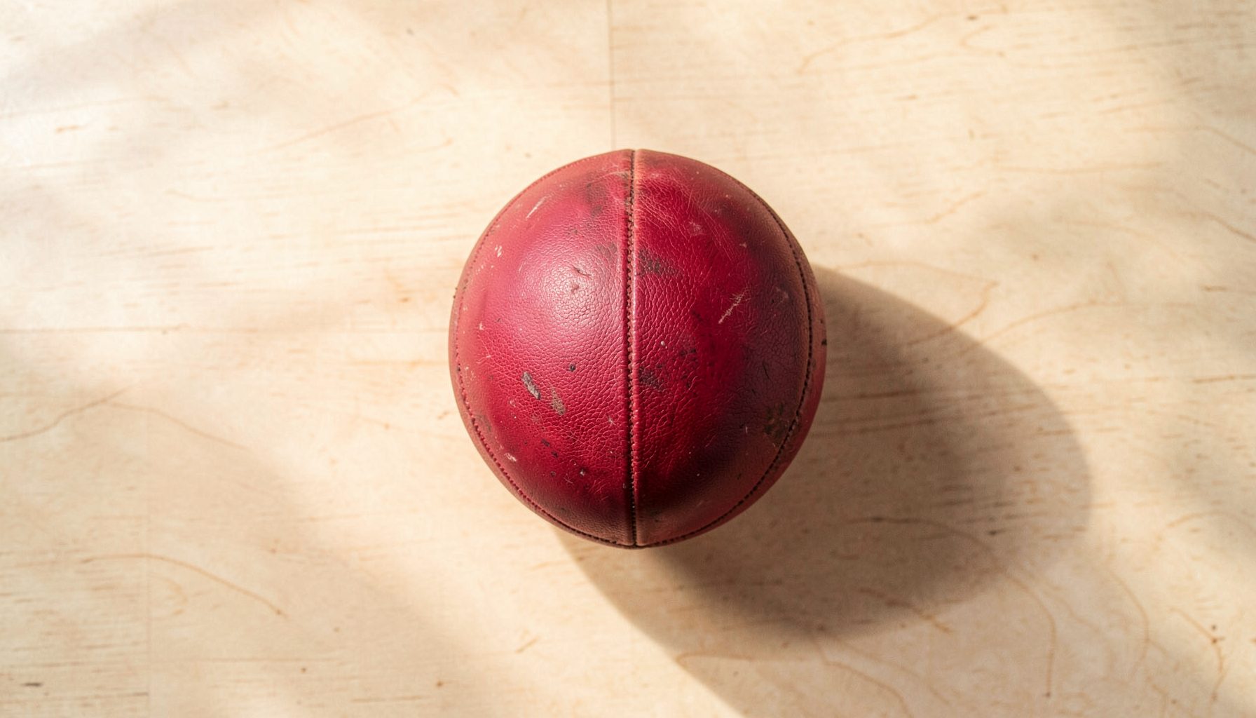 Overhead flat-lay photograph of a worn leather Rugby Fives ball resting on the pale wooden floor of a court, with soft shadows stretching across the grain; warm cream and deep maroon tones dominate the scene.