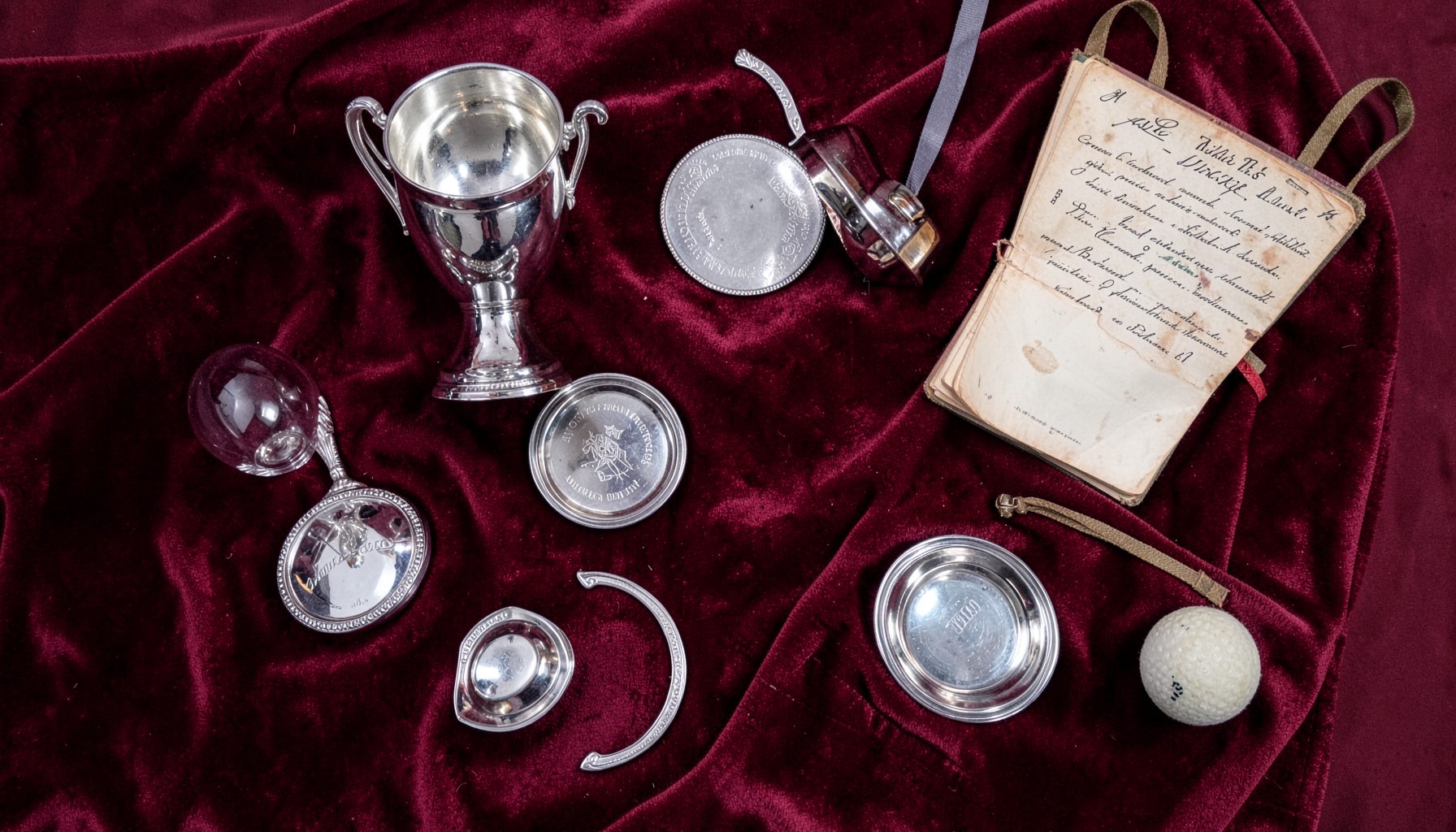 Overhead flat-lay of silver trophies and engraved plates arranged on rich burgundy velvet, with a vintage scorebook and a fives ball, deep maroon and silver tones