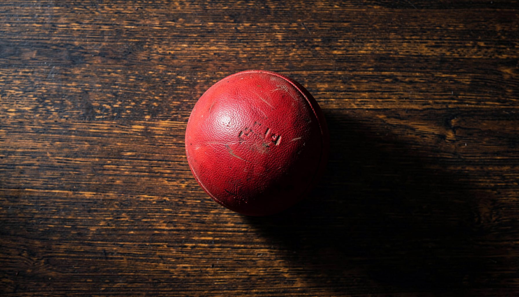Overhead flat-lay of a worn leather Rugby Fives ball resting on the dark wooden floor of a court, with soft side-lighting casting long shadows across the grain; deep browns, warm amber, and muted charcoal tones.