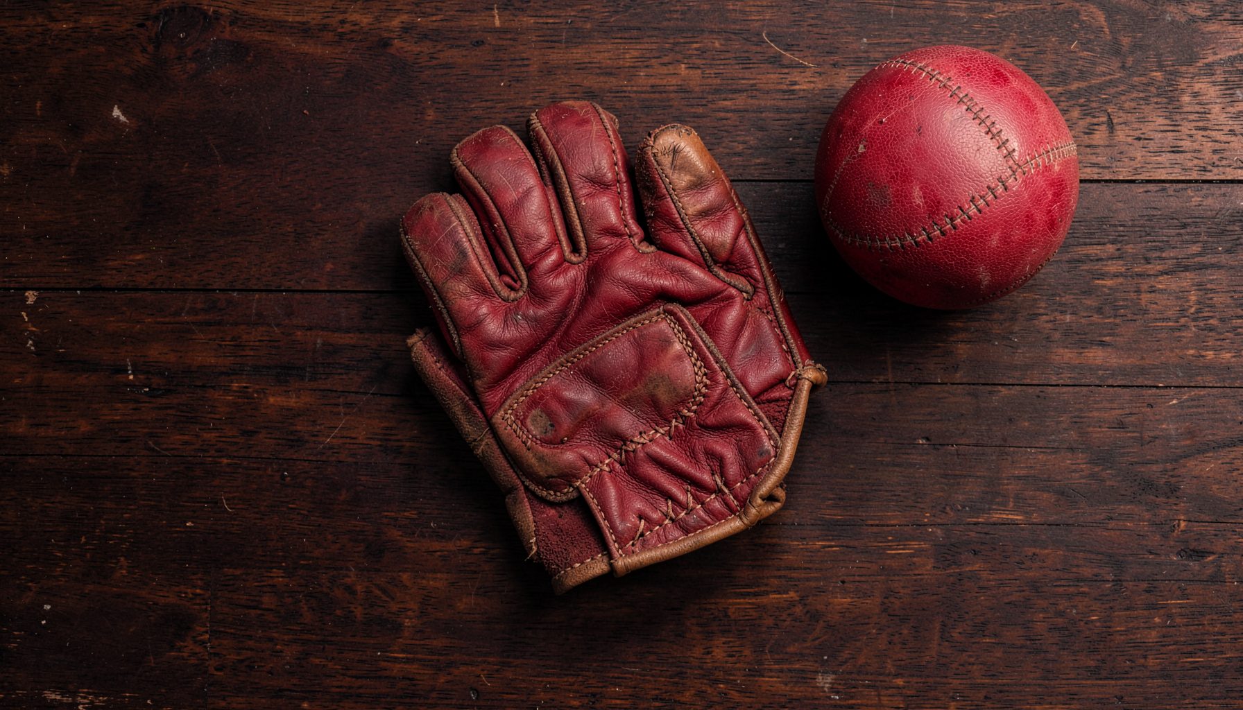 Overhead flat-lay photograph of a worn leather Rugby Fives glove resting on a dark wooden court floor, with a hand-stitched ball nearby; deep browns, burgundy shadows, and warm cream highlights evoke a vintage sporting atmosphere.