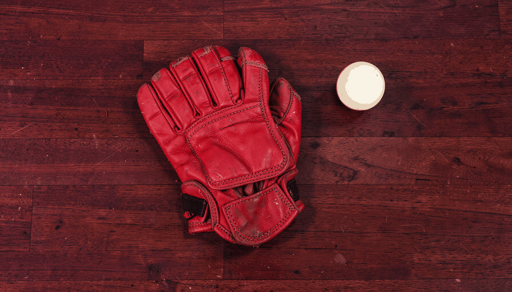 Overhead flat-lay photograph of a worn leather Rugby Fives glove resting on a dark wooden court floor, with a single white ball nearby; deep maroon and warm brown tones dominate, with soft cream highlights.